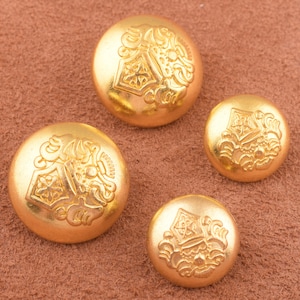 Puede incluir: Cuatro botones de metal de color dorado con diseños intrincados. Los botones tienen un patrón adornado en relieve y un acabado liso y brillante.