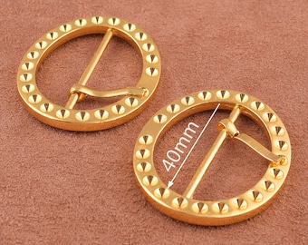 May include: Two gold-toned metal belt buckles with a circular design and a studded edge. The buckle on the right has a measurement of 40mm.