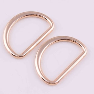 Puede incluir: Dos anillos en D de metal dorado. Los anillos son lisos y tienen un acabado pulido.