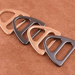 Op de afbeelding: Vier metalen D-ringgespen in diverse metallic afwerkingen: roségoud, gunmetal en goud. De gespen zijn diagonaal geplaatst op een bruin, getextureerd oppervlak. Geschikt voor leerbewerking.
