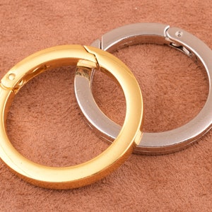 Op de afbeelding: Twee sleutelhangers, een gouden en een zilveren, worden getoond op een bruine suède ondergrond. De sleutelhangers zijn beide rond en hebben een veerbelaste sluiting.