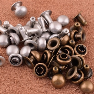 May include: A collection of silver and brass metal rivets, some with a domed top and others with a flat top. The rivets are arranged in a pile on a brown leather surface.