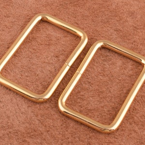 May include: Two gold-colored metal rectangular rings or loops. The rings are smooth and have rounded edges.
