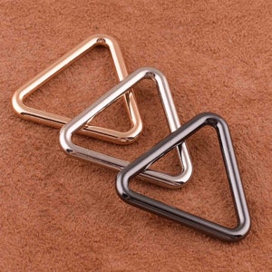 Puede incluir: Tres anillos triangulares metálicos en oro rosa, plata y gris plomo. Los anillos están dispuestos en diagonal sobre una superficie marrón texturizada. Estos componentes de hardware son probablemente para la fabricación de bolsos u otras manualidades.