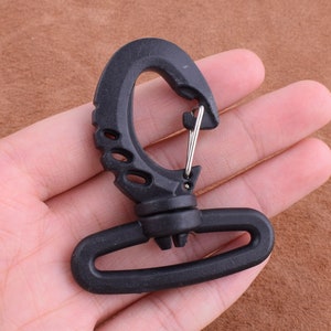 May include: A black plastic swivel hook with a metal spring clip. The hook has a curved design with several oval cutouts and a rotating base. The hook is held in a hand.