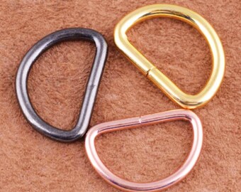 May include: Three D-shaped metal rings in different finishes: black, gold, and rose gold. The rings are on a brown surface.