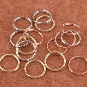 Pode incluir: Uma coleção de chaveiros sortidas em tons prata, dourado e cobre. Os chaveiros estão dispostos em uma pilha sobre uma superfície de couro marrom.
