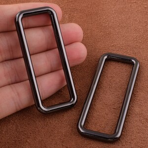 Peut inclure: Deux anneaux rectangulaires en métal noir, également appelés anneaux en D, sont présentés sur une surface en cuir brun. Les anneaux mesurent environ 5 cm de long et 2,5 cm de large.