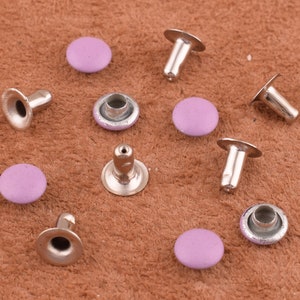 May include: Close-up of several silver-colored rivets with light purple, circular tops. The rivets are scattered on a brown, textured surface. The rivets are designed for use in leathercraft or similar projects.