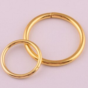 May include: Two gold-coloured metal rings, one larger than the other, are shown against a light pink background. The rings are smooth and have a shiny finish.