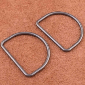 May include: Two silver metal D-rings, each with a rounded top and a flat bottom. The rings are on a brown suede surface.