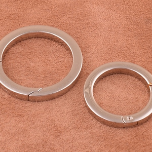 Puede incluir: Dos llaveros de metal plateado con cierre de resorte. Los llaveros están sobre una superficie de gamuza marrón.