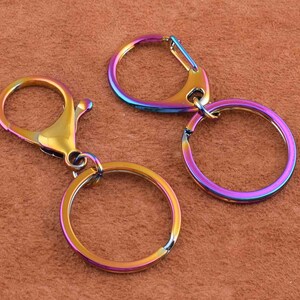Peut inclure: Trois porte-clés irisés avec une finition arc-en-ciel. L'un a un mousqueton, et les deux autres ont des anneaux fendus. Les porte-clés sont en métal et ont une surface lisse et polie. Les couleurs passent de l'or au bleu et au violet.