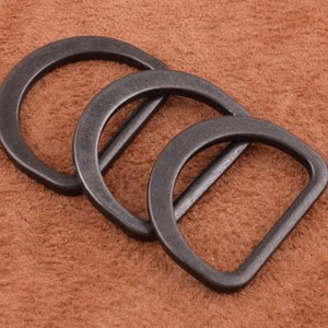 May include: Three black metal D-rings, also known as belt loops, are arranged in a stack on a brown suede surface.