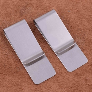 Puede incluir: Dos clips para dinero de metal plateado. Los clips son rectangulares con esquinas redondeadas y una ligera curva en el medio. Los clips están hechos de metal liso y cepillado.