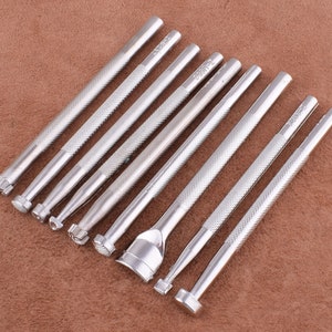 May include: Set of nine silver metal leather craft tools with different shaped tips. The tools are arranged in a row on a brown textured surface.