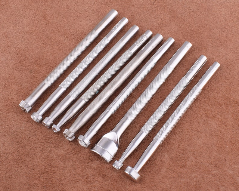 May include: Set of nine silver metal leather craft tools with different shaped tips. The tools are arranged in a row on a brown textured surface.