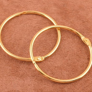 Puede incluir: Dos anillos de encuadernación de metal dorado, uno ligeramente más grande que el otro, están apilados uno encima del otro. Los anillos son lisos y brillantes.