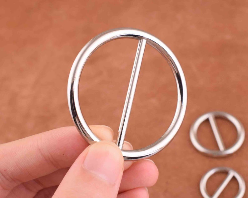 Puede incluir: Hebilla de cintur&oacute;n de metal plateado de forma redonda con una barra recta en el centro.