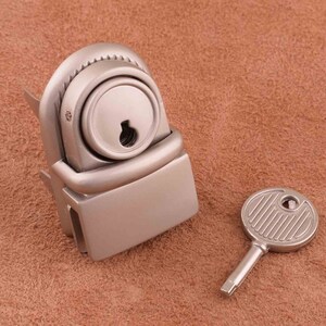 Puede incluir: Una cerradura de metal plateada con un ojo de cerradura y una llave a juego. La cerradura tiene una forma rectangular con una parte superior redondeada y un pequeño ojo de cerradura circular. La llave es redonda con un diseño acanalado.