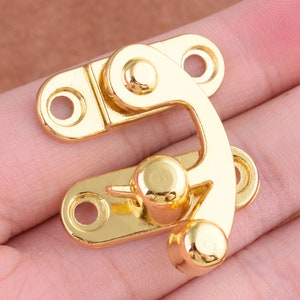 Puede incluir: Cierre de metal dorado con un pestillo giratorio y dos agujeros para sujetar a la tela. El cierre tiene un diseño decorativo y es adecuado para proyectos de artesanía o costura.