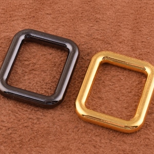 May include: Two metal square rings, one black and one gold, on a brown suede surface.