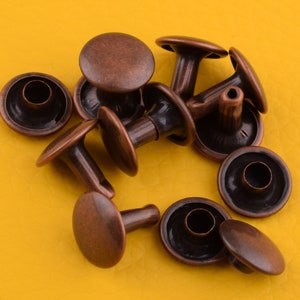 May include: A group of copper-colored metal rivets with a round head and a post. The rivets are arranged in a pile on a yellow surface.