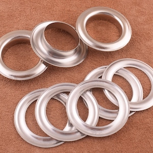 May include: A set of silver metal eyelets for crafts and leatherwork. The eyelets are in various sizes and are arranged on a brown surface.