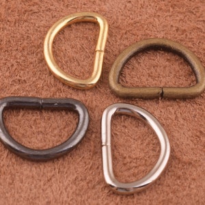 May include: Four D-rings in different finishes: gold, brass, silver, and black. The rings are all the same size and shape, and they are lying on a brown leather surface.