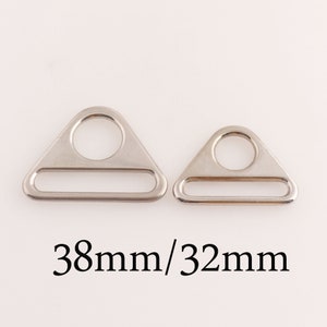 Pode incluir: Dois anéis de metal em forma de triângulo prateados com uma abertura retangular e um furo redondo no centro. O anel maior mede 38 mm e o menor 32 mm.