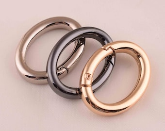 May include: Three oval-shaped metal clasps in silver, gunmetal, and rose gold. The clasps are open, showcasing their spring-loaded mechanisms. These hardware components are likely used for bags, jewelry, or other crafting projects.