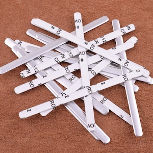 May include: A pile of silver metal nose wire strips for face masks. The strips are about 5 inches long and have a white adhesive backing with black printed text that includes "RO", "512", "CR", "WN", "OV", and "#" symbols.