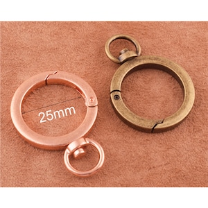 Pode incluir: Dois chaveiros de metal, um em ouro rosa e outro em latão antigo. O chaveiro de ouro rosa tem um diâmetro de 25 mm e um pequeno laço na parte inferior. Ambos os chaveiros têm um fecho de mola.