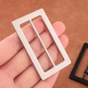 May include: A silver metal belt buckle with a rectangular shape and two slots for a belt strap.