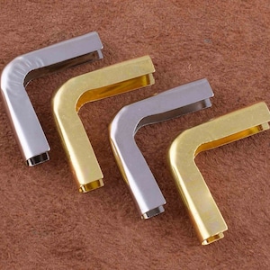 May include: Four metal corner protectors in different finishes: silver, gold, and two shades of gold. The corner protectors are L-shaped and have a smooth, polished finish.