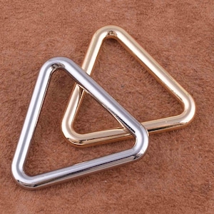 Pode incluir: Dois anéis de metal em forma de triângulo, um prateado e outro dourado, são mostrados em uma superfície marrom. Os anéis estão entrelaçados.