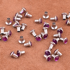 May include: A collection of silver-toned metal studs with purple rhinestones. The studs are scattered on a brown surface.