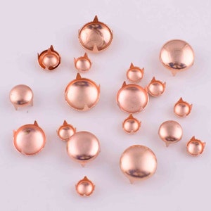 Puede incluir: Surtido de tachuelas de metal color oro rosa en varios tamaños. Las tachuelas tienen una parte superior abovedada y cuatro puntas.