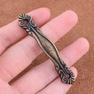May include: An antique bronze-colored cabinet pull with an ornate, symmetrical design. The pull has a curved center and decorative ends with circular holes for mounting. The hardware is held in a hand, showcasing its size and detail.
