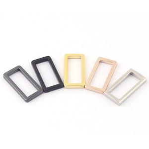 May include: Five metal rectangular rings in different finishes: black, silver, gold, rose gold, and gunmetal. The rings are all the same size and shape.
