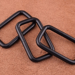 May include: Three black metal D-rings, each with a different size, are arranged on a brown textured surface.