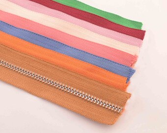 Puede incluir: Surtido de cintas de cremallera en varios colores, incluyendo verde, rojo, rosa, beige, naranja y marrón. Las cintas de cremallera están dispuestas en una pila, con la cinta marrón en la parte inferior y la cinta verde en la parte superior. Las cintas de cremallera tienen una cremallera de metal plateado.