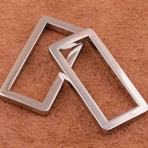 May include: Two silver metal rectangular belt buckle loops. The loops are interlocking and have a smooth, polished finish.