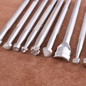 May include: A set of ten silver metal leather craft tools with different shaped tips. The tools are arranged in a row on a brown surface.