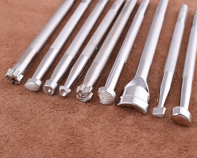 May include: A set of ten silver metal leather craft tools with different shaped tips. The tools are arranged in a row on a brown surface.