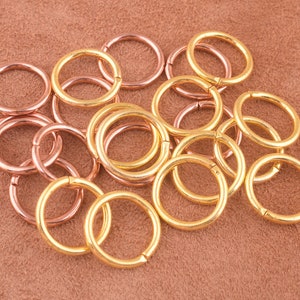 May include: A collection of gold and rose gold jump rings. The jump rings are small and circular, and they are arranged in a pile on a brown surface.