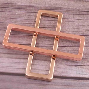 May include: Rose gold metal buckle with a rectangular shape and a cross-shaped design. The buckle has two openings for straps.