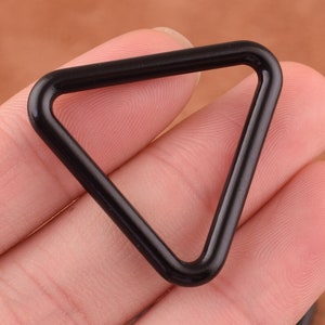 Può includere: Un anello di metallo triangolare nero. L'anello è liscio e ha una finitura lucida.