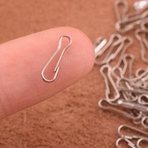 Pode incluir: Um fecho de mosquetão de metal prateado, também conhecido como fecho de garra de lagosta, é mostrado em um dedo. O fecho é pequeno e tem um acabamento liso e brilhante.