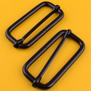 Peut inclure: Deux boucles coulissantes en métal noir. Les boucles sont rectangulaires avec un bord arrondi et une seule barre horizontale centrée.